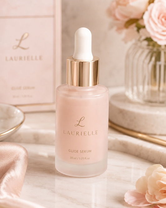 Laurielle Glide Serum in a frosted glass bottle with a white dropper cap and gold collar, set on a marble surface with a pink product box and soft pink flowers in the background.